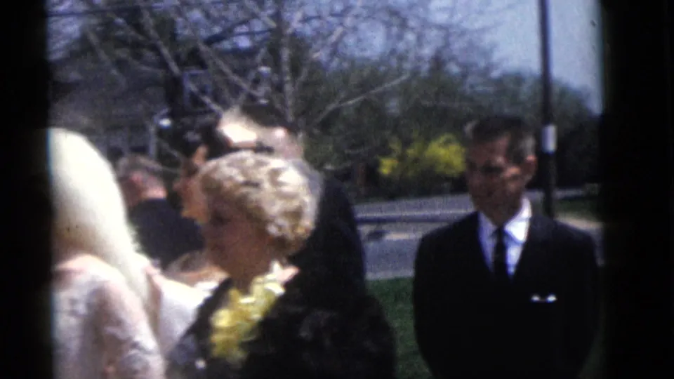 1960 Camden wedding home-movie frame with the bride partly passing the camera, guests nearby, and a suited attendant on the lawn.