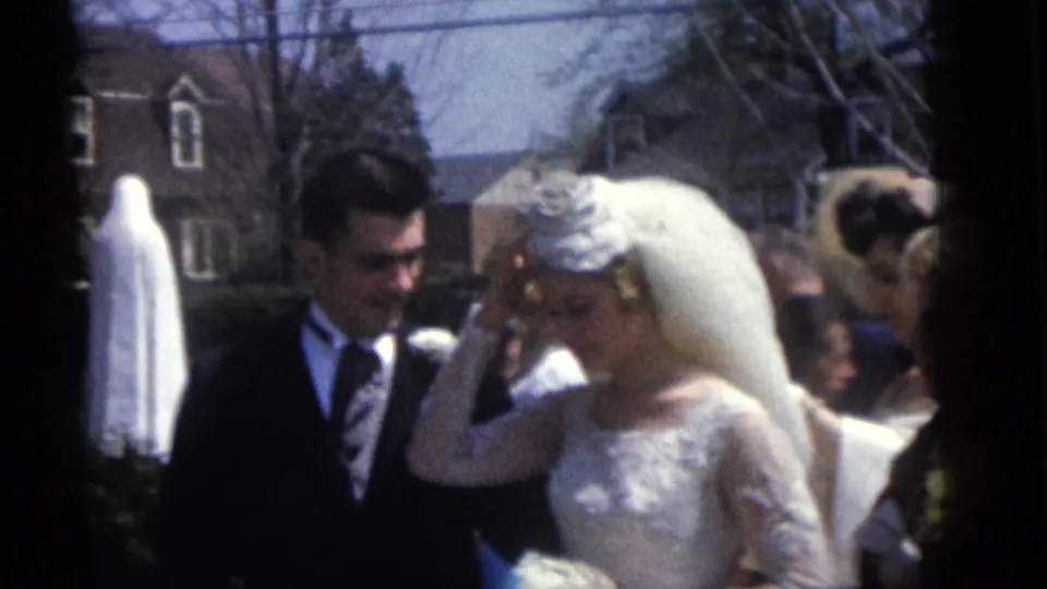 1960 Camden wedding frame showing the bride adjusting her headpiece beside the groom, with guests and a white statue behind them.