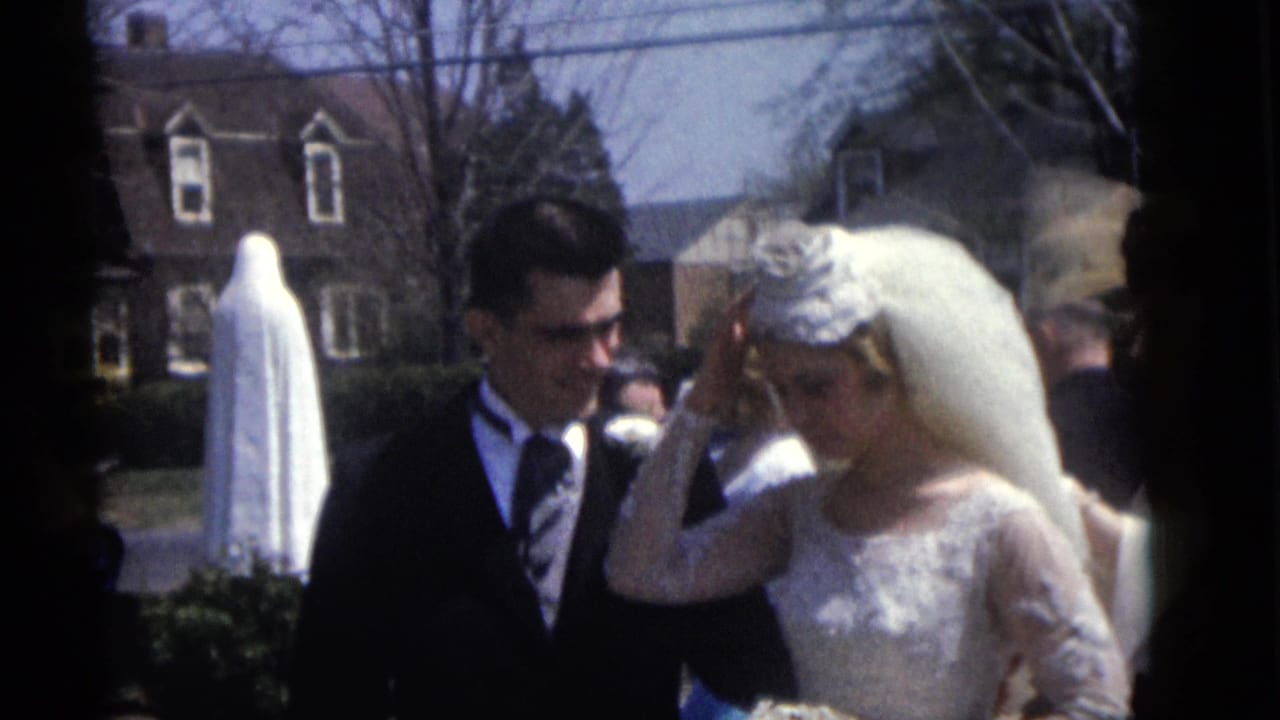 Bride adjusts her veil while the groom stands beside her outdoors near a white-robed statue.