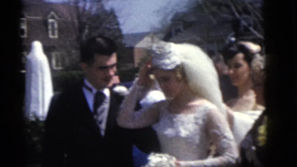 The bride raises her hand to her veil while the groom stands close beside her in the Camden wedding scene, with guests and a statue in back.