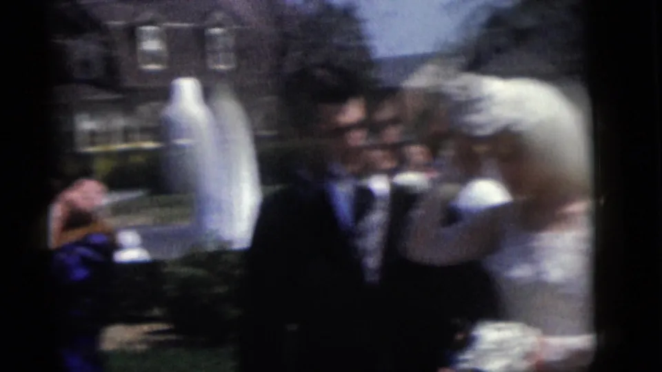 A motion-blurred 8mm frame shows the bride and groom shifting position outdoors in the 1960 Camden wedding footage, with the statue still visible.