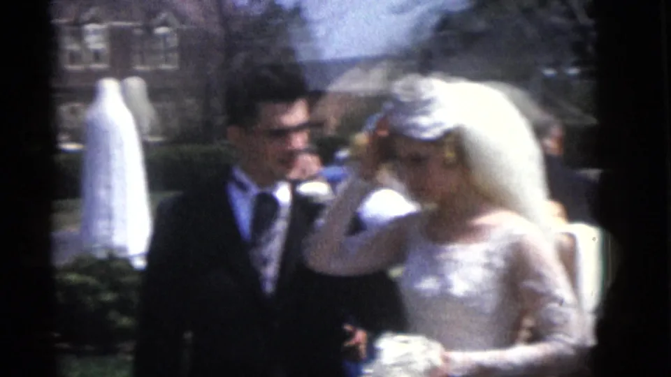 The bride and groom remain side by side in the 1960 Camden wedding home movie as she holds her veil, with a white-robed statue in the background.