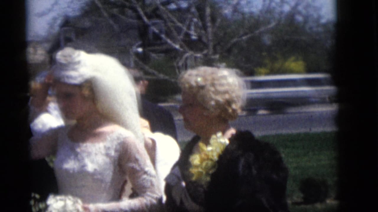 In 1960 Camden, New Jersey, a bride in a white lace gown and long veil stands near wedding guests in hats outdoors.