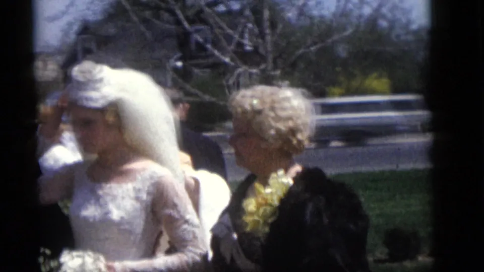 In 1960 Camden, New Jersey, a bride in a white lace gown and long veil stands near wedding guests in hats outdoors.
