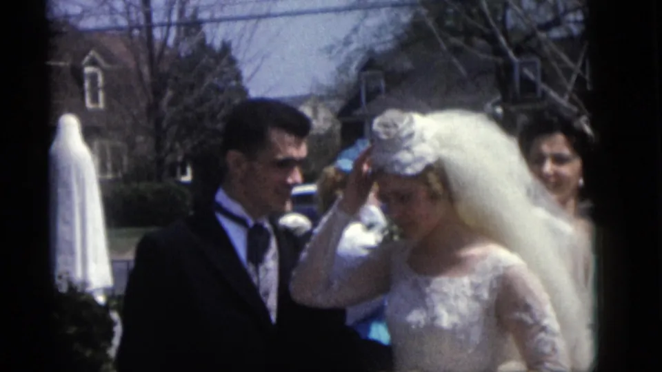 A groom in a dark suit stands close to the bride as she touches her veil in this Camden wedding home-movie frame.