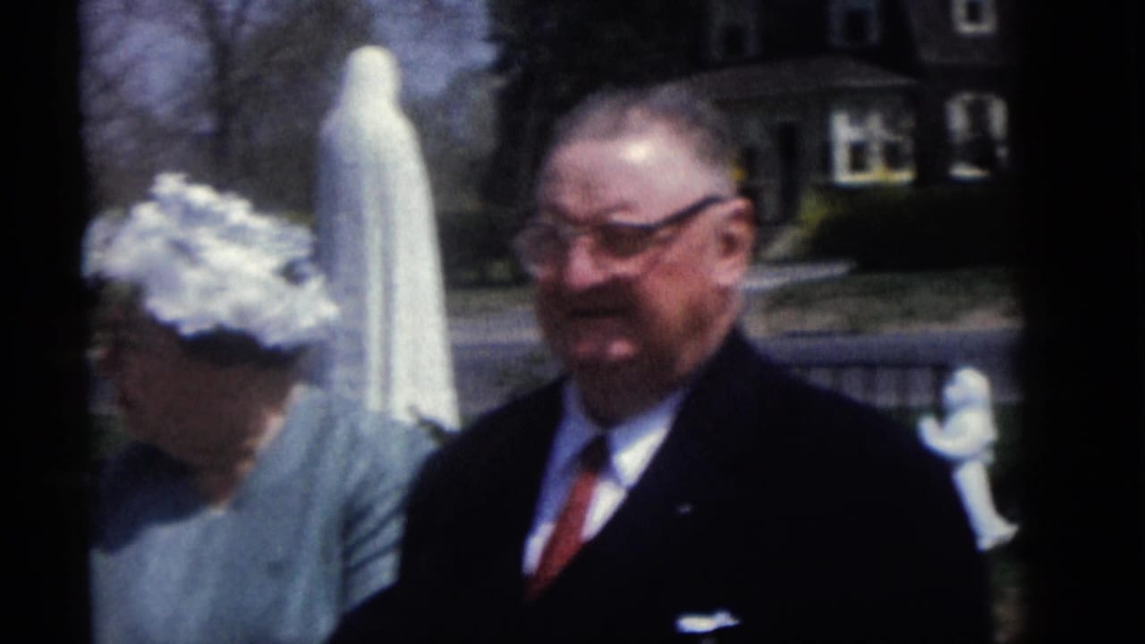 Man in a dark suit and woman in a white hat stand beside a white robed statue in a suburban yard.