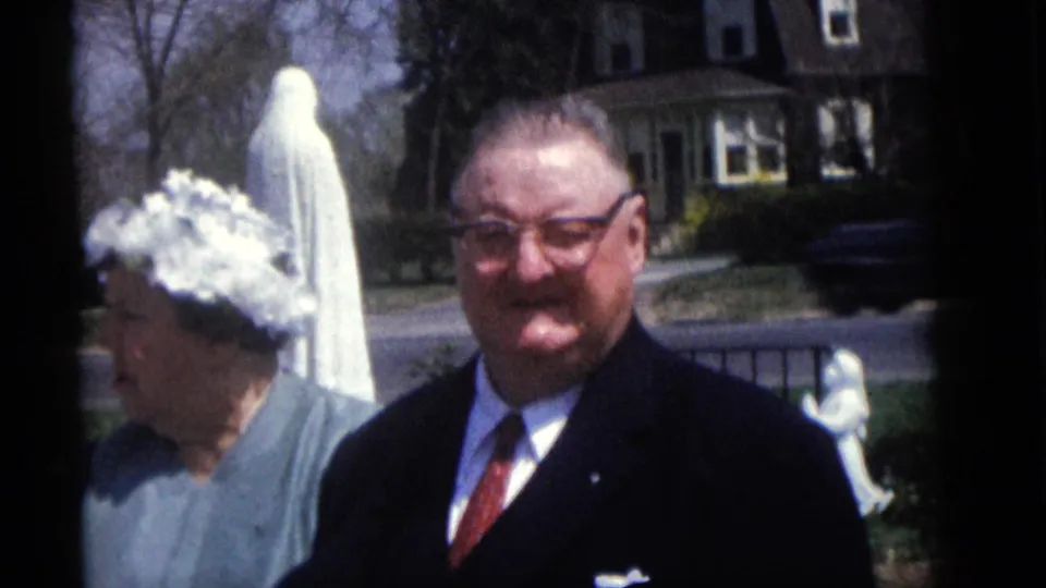 A suited man looks toward the camera in Camden, New Jersey, with a woman, white yard statue, fence, and passing street traffic nearby.