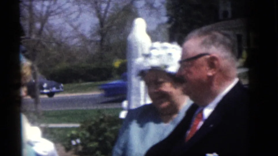 The Camden home-movie frame shows the suited man turned toward the woman beside a white yard statue, with cars and houses in the background.
