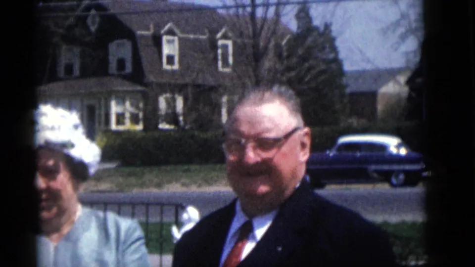 A smiling suited man with glasses stands beside a woman in a white hat in the Camden, New Jersey wedding home movie.