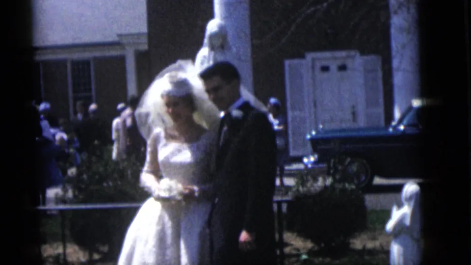 Bride with veil and bouquet stands next to the groom in a dark suit outside during the 1960 Camden wedding footage.