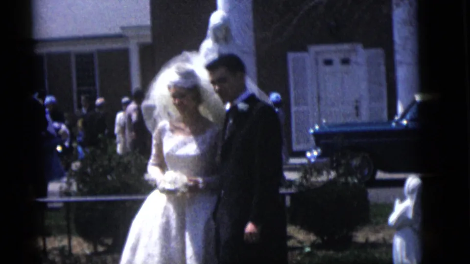 The bride and groom remain outdoors near white doors and garden statues, with guests and a dark car behind them.