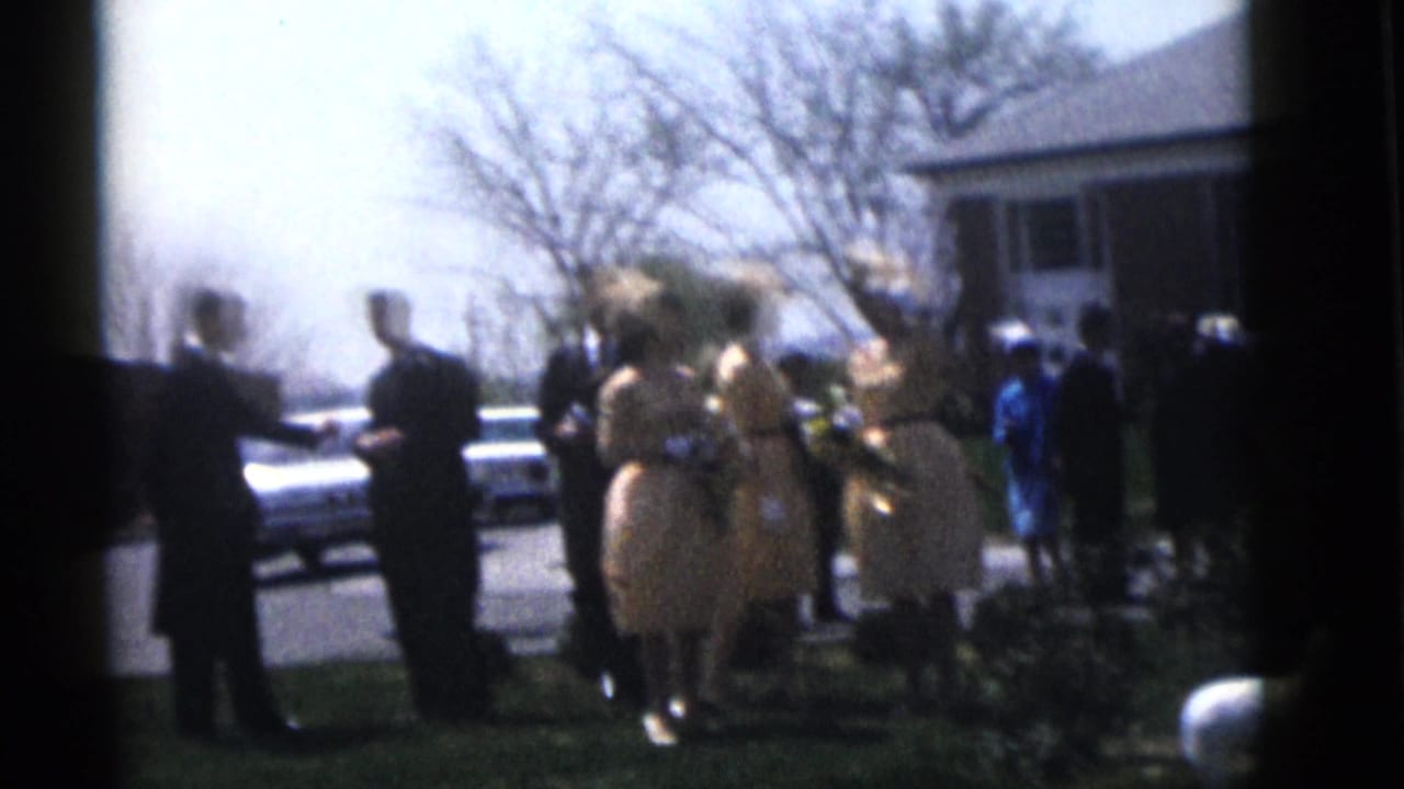 1960 Camden wedding guests in formal clothing gather outside a brick building, with cars and bare trees in the background.