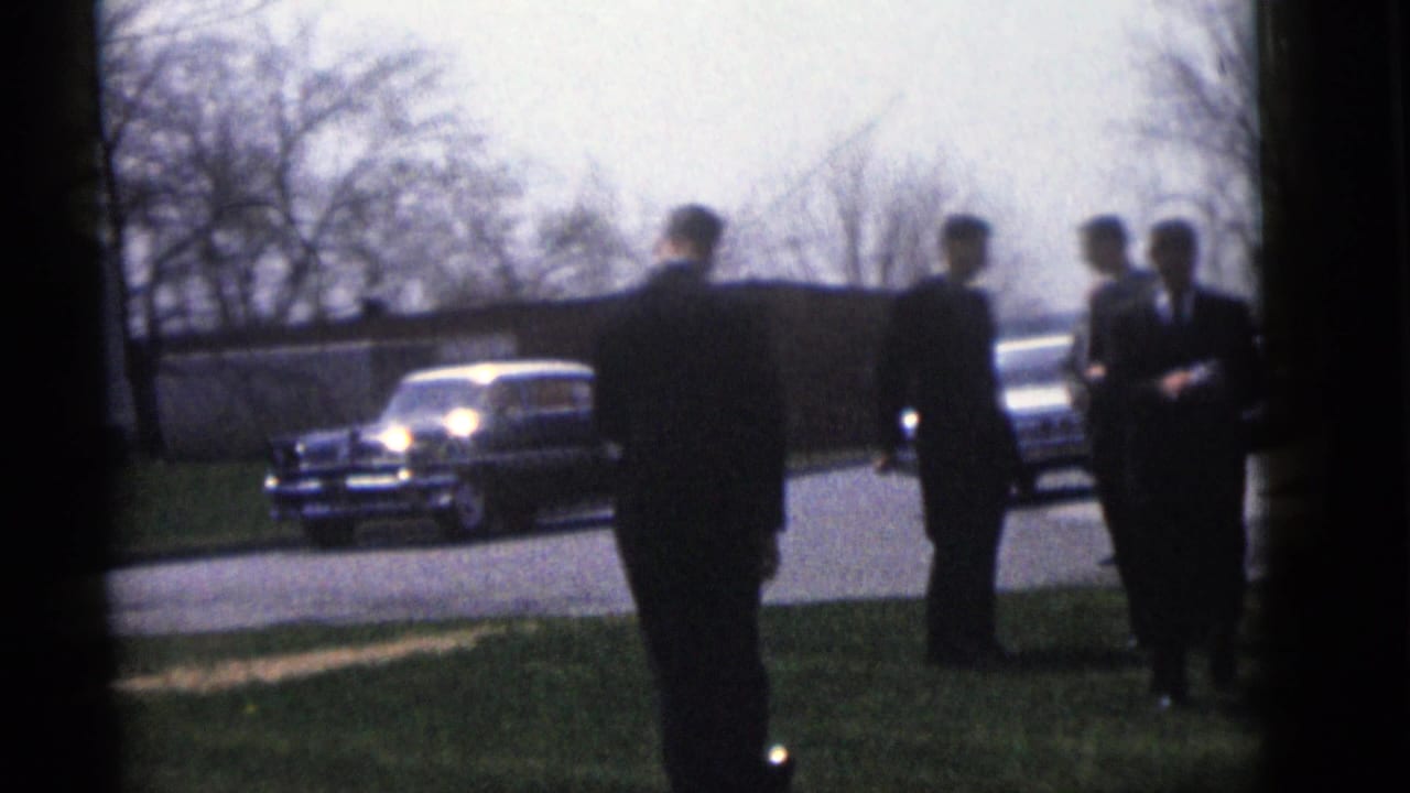 Men in formal suits stand outside during the Camden wedding gathering, with mid-century cars parked near the road.
