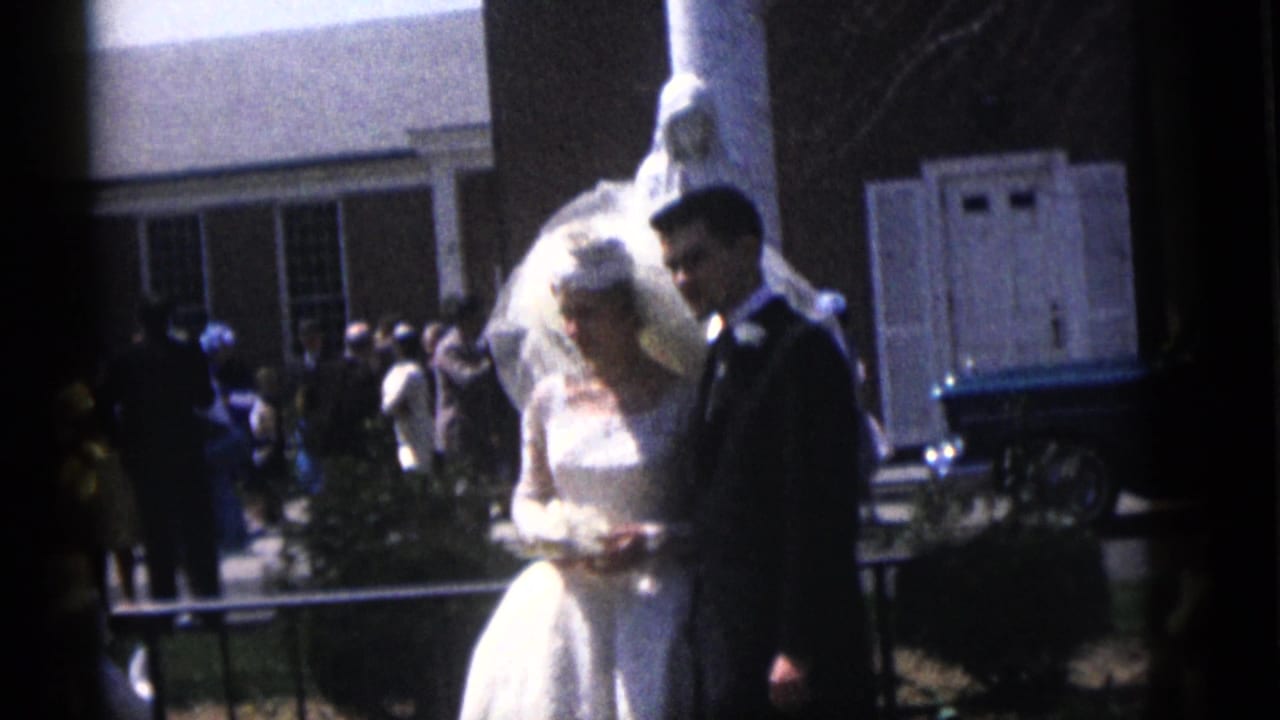 The bride and groom stand together at the Camden wedding, with guests, a brick building, and a white lamb figure behind them.
