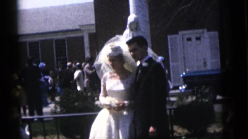 The bride and groom stand together at the Camden wedding, with guests, a brick building, and a white lamb figure behind them.