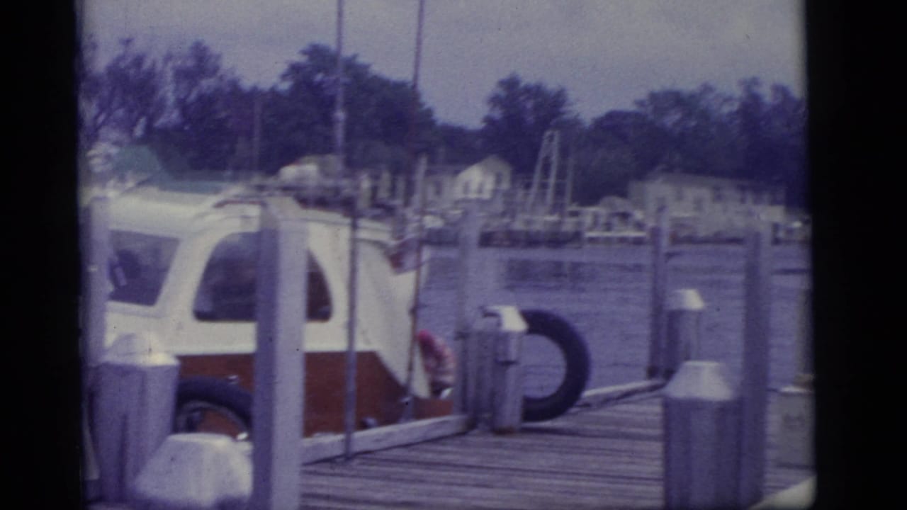 White and red boat moored beside a wooden dock with water and houses behind it.