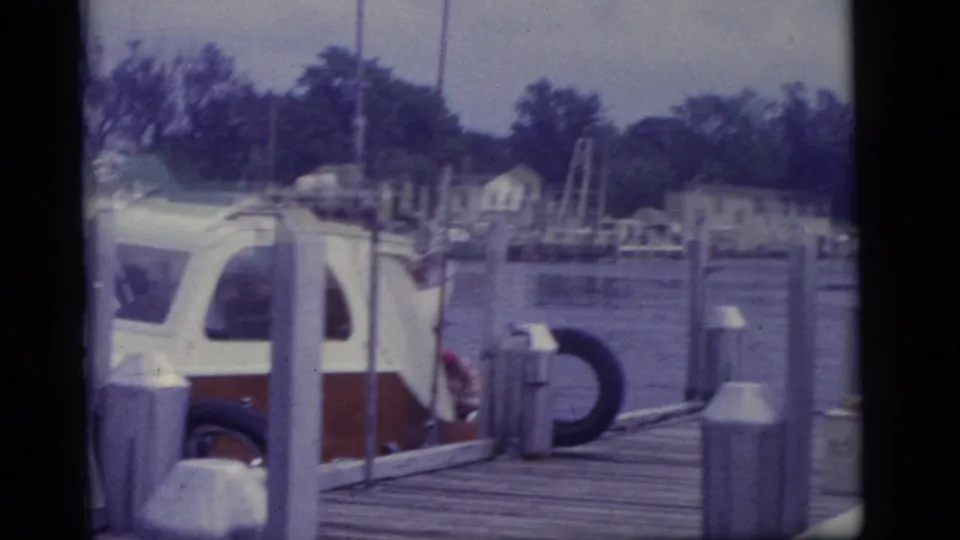 1960 Camden home-movie frame showing a small white and red boat near dock posts with calm water and distant houses.