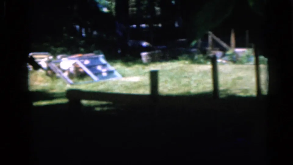 1960 Camden, New Jersey home-movie frame showing a grassy yard, fence posts, shrubs, and a small ramp or toy structure.
