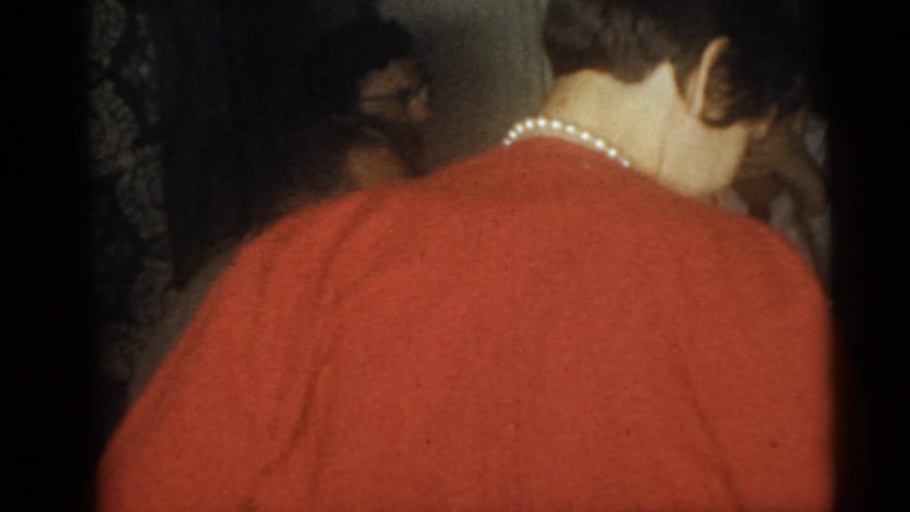 1960 Camden home-movie frame with a woman in a red sweater and pearl necklace among people inside a private room.