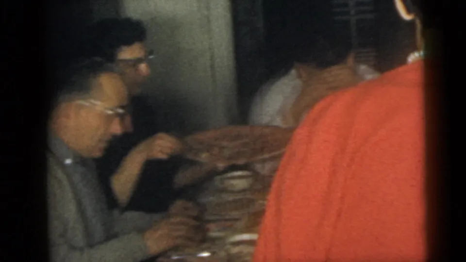 1960 Camden home-movie frame showing adults seated and standing around a table during an indoor family gathering.