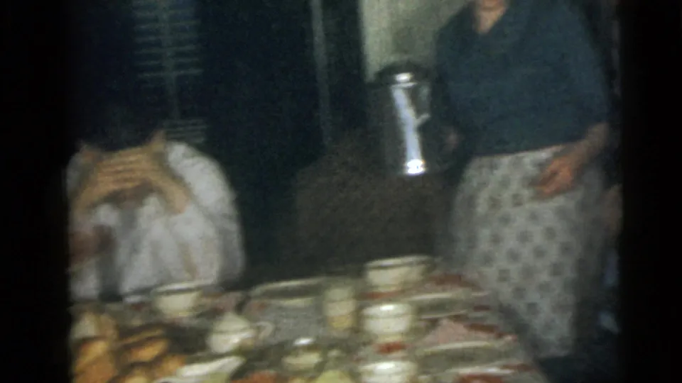 1960 Camden home-movie frame showing a table set with dishes and food while people stand nearby in a private interior.