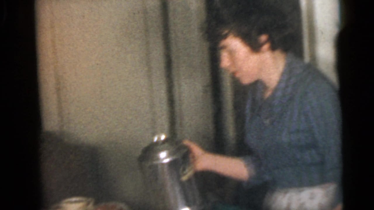 Woman in a blue patterned dress pours from a metal pitcher inside a dining room.