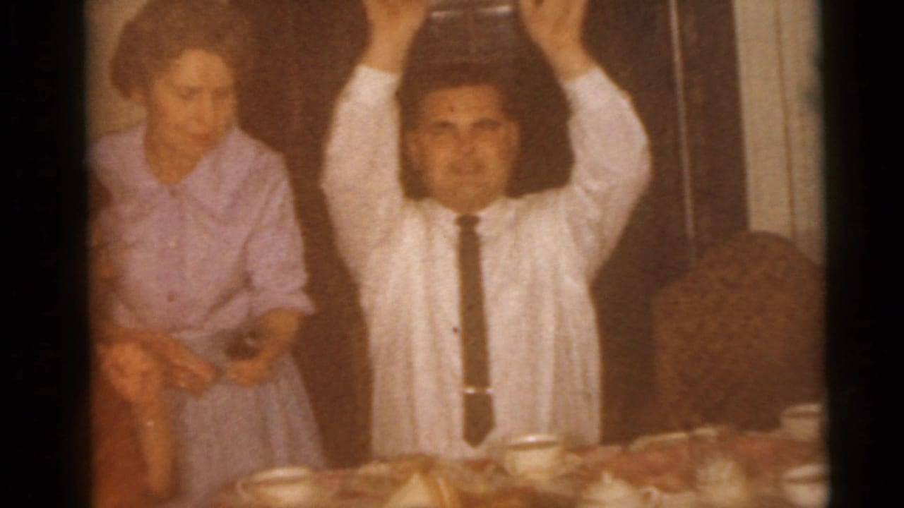 Adults sit at a dining table while a man in a white shirt raises both hands.