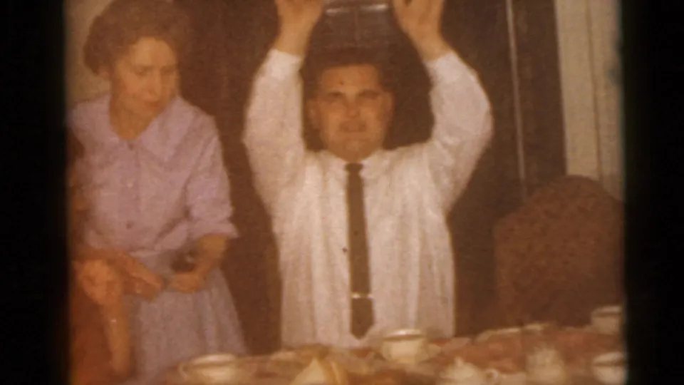 A 1960 Camden home-movie frame shows adults at a dining table, with a man in a white shirt and tie raising both hands.
