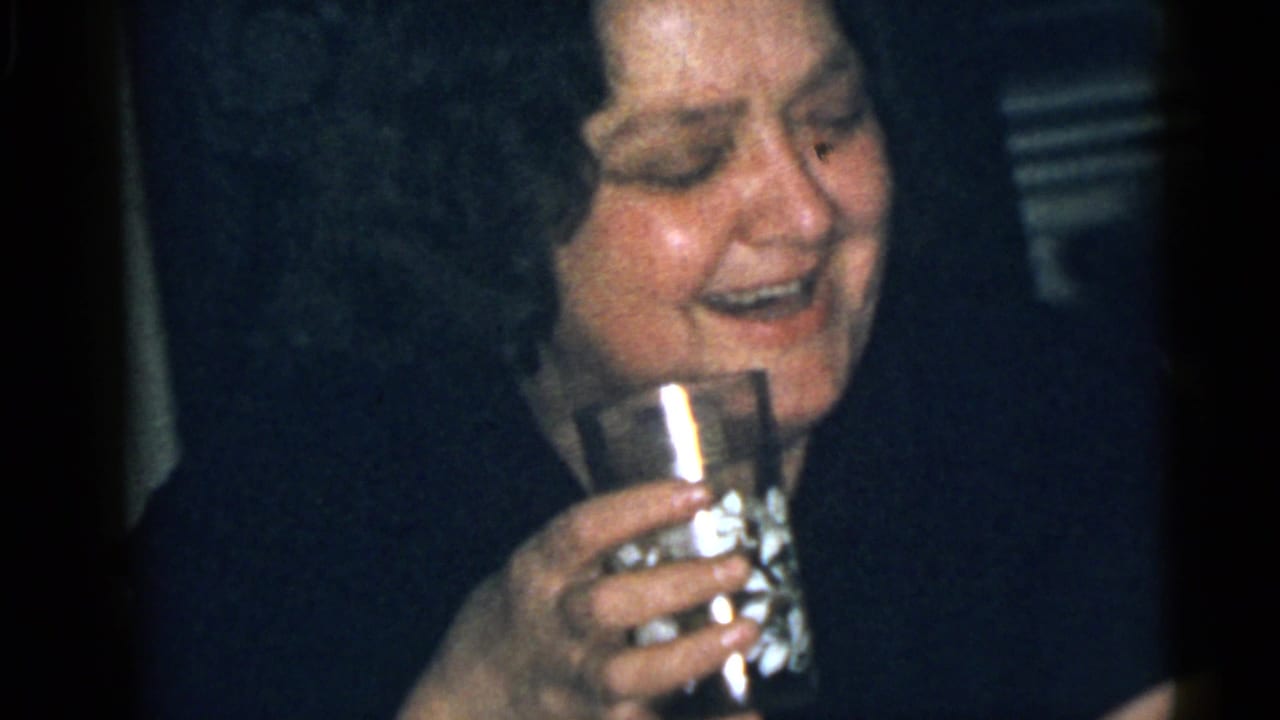 Adult woman in a dark top smiles while holding a drinking glass indoors.