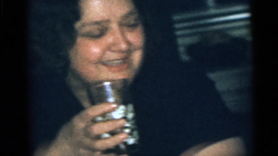 The woman continues laughing with a drinking glass in hand, framed closely in soft 8mm indoor light.