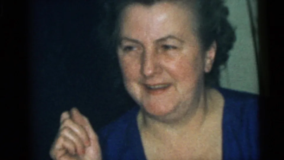 An older woman in a blue top appears in the Camden home-movie scene, looking aside during the indoor gathering.