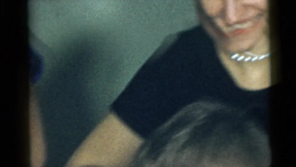 A partially framed adult wearing a dark top and light necklace smiles near the edge of the 1960 Camden home-movie frame.