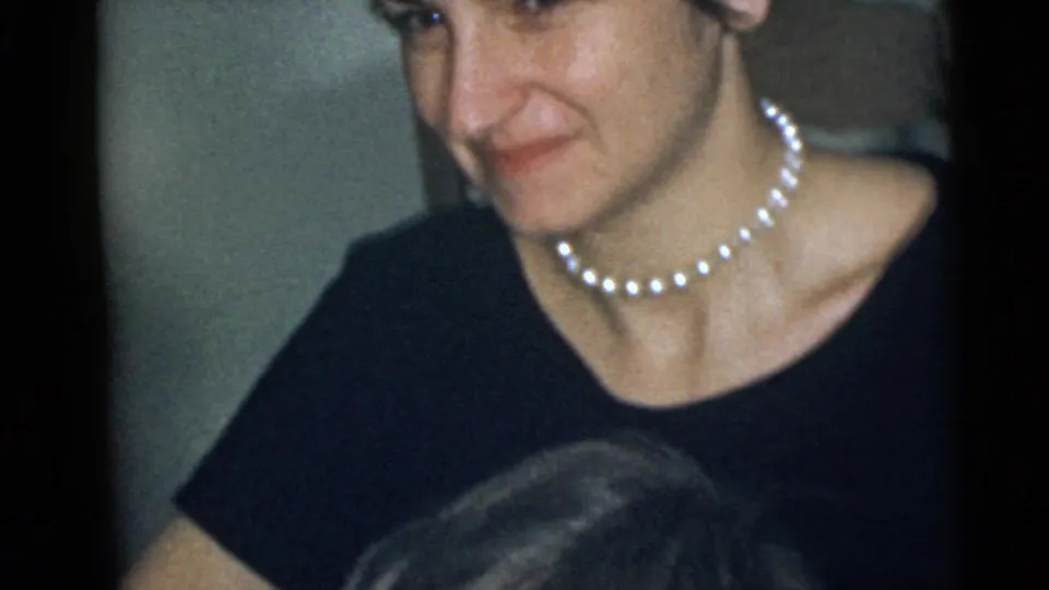 Adult woman looks downward in a close 8mm frame, with a pearl necklace, dark top, and indoor background visible.