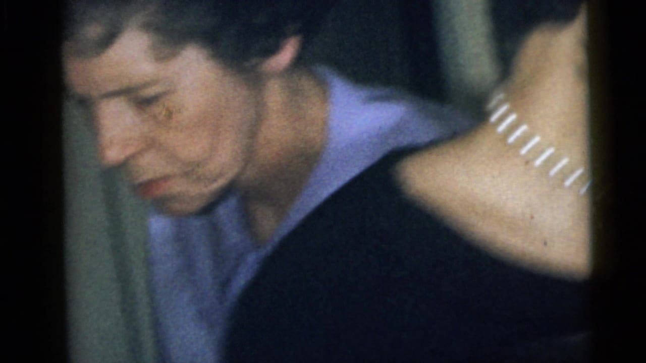 1960 Camden home-movie frame showing an adult woman in a light purple collared shirt looking down indoors, with another person partly visible.