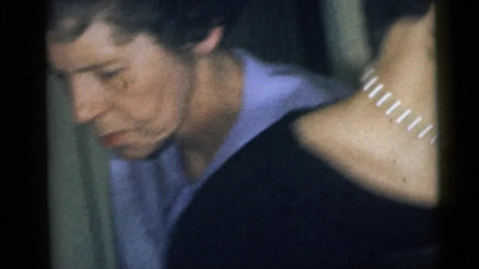 1960 Camden home-movie frame showing an adult woman in a light purple collared shirt looking down indoors, with another person partly visible.