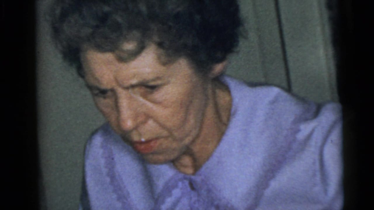 Adult woman in a light purple collared shirt looks down beside a plain interior wall.