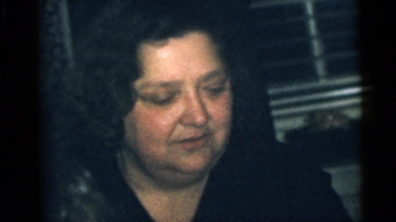 1960 Camden home-movie frame of a dark-haired woman seated in a private interior with window blinds behind her.