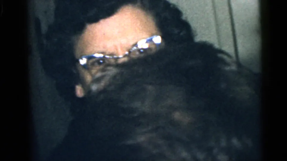 1960 Camden home-movie frame showing a woman with glasses partly obscured by a close foreground figure in a private room.