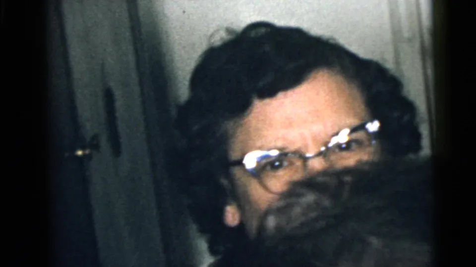 1960 Camden home-movie frame of a woman with glasses in a dim interior, partly obscured by someone close to the camera.