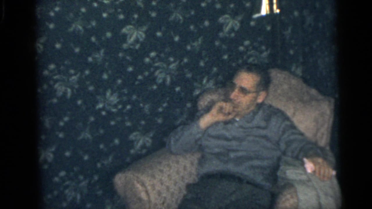 In a Camden, New Jersey 1960 home-movie frame, an adult man reclines in an armchair against floral wallpaper.