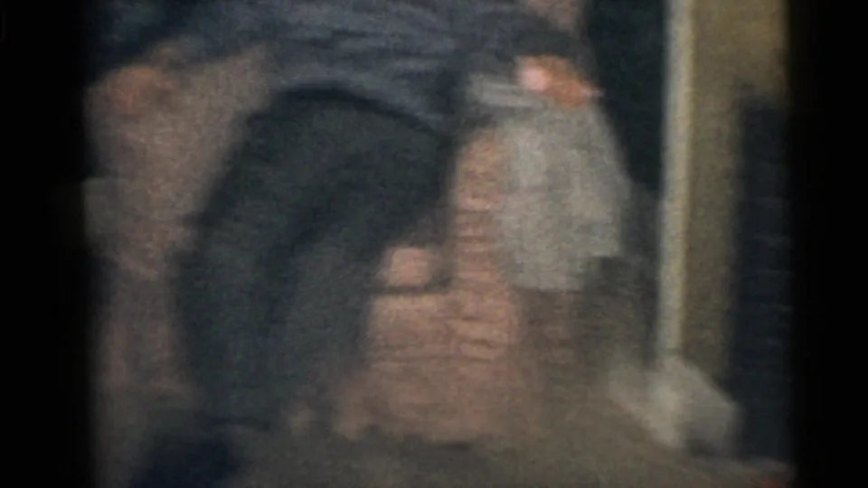 A soft and blurred 1960 home-movie frame appears to show an adult figure seated indoors near upholstered furniture.