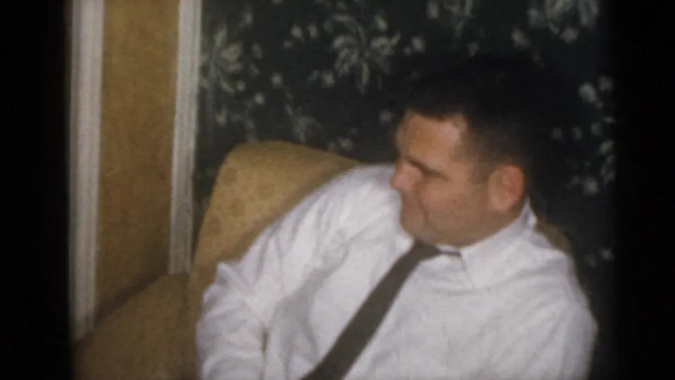 An adult man in a white dress shirt and dark tie sits in an armchair in the Camden 1960 home-movie interior.