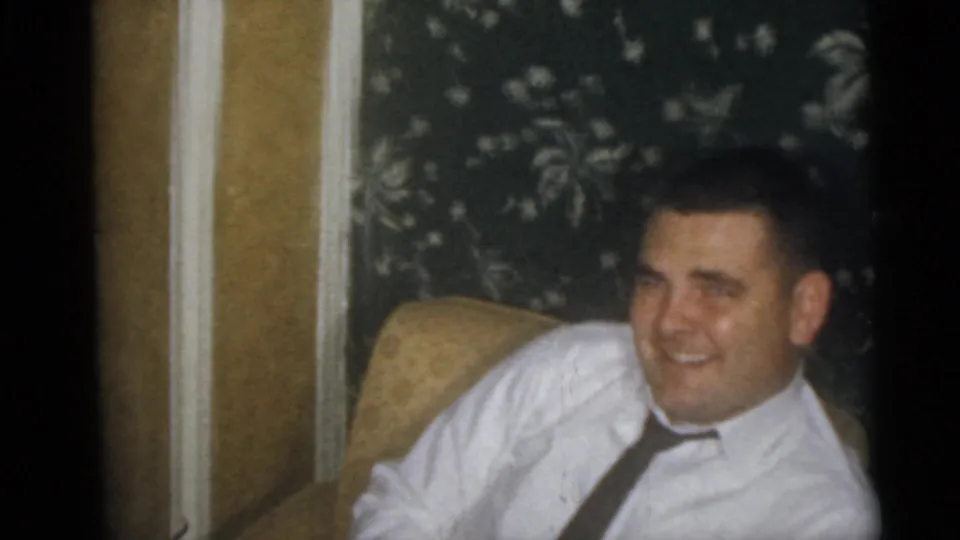 An adult man in a white shirt and tie smiles from an armchair in a Camden, New Jersey 1960 home-movie interior.