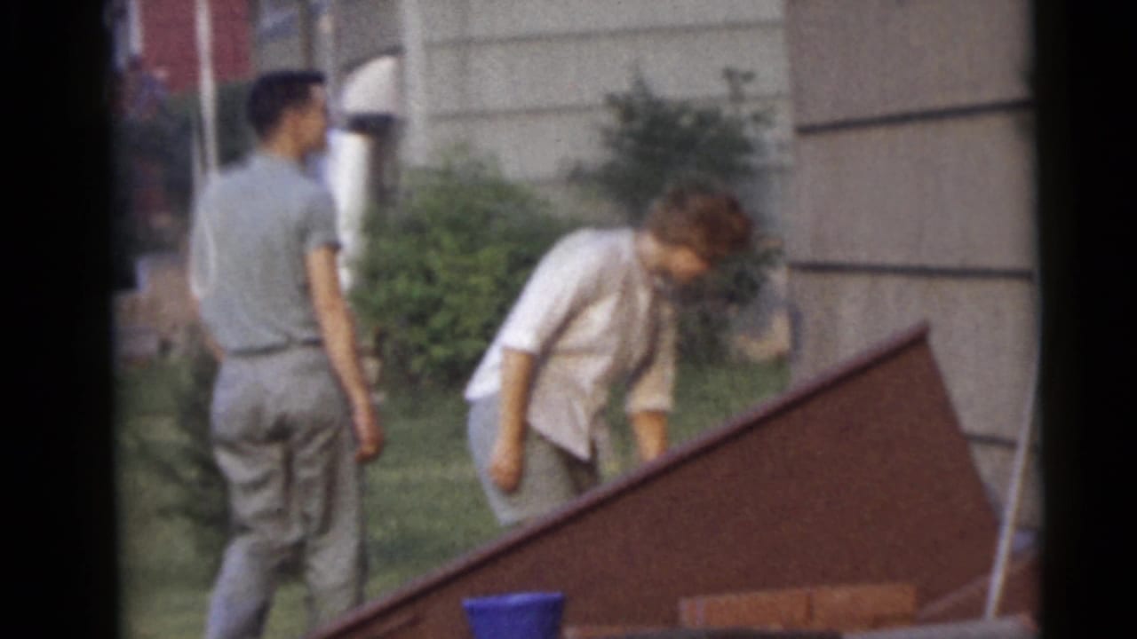 In Camden, New Jersey home-movie footage from 1960, a man and woman walk through a grassy backyard beside shrubs and house siding.