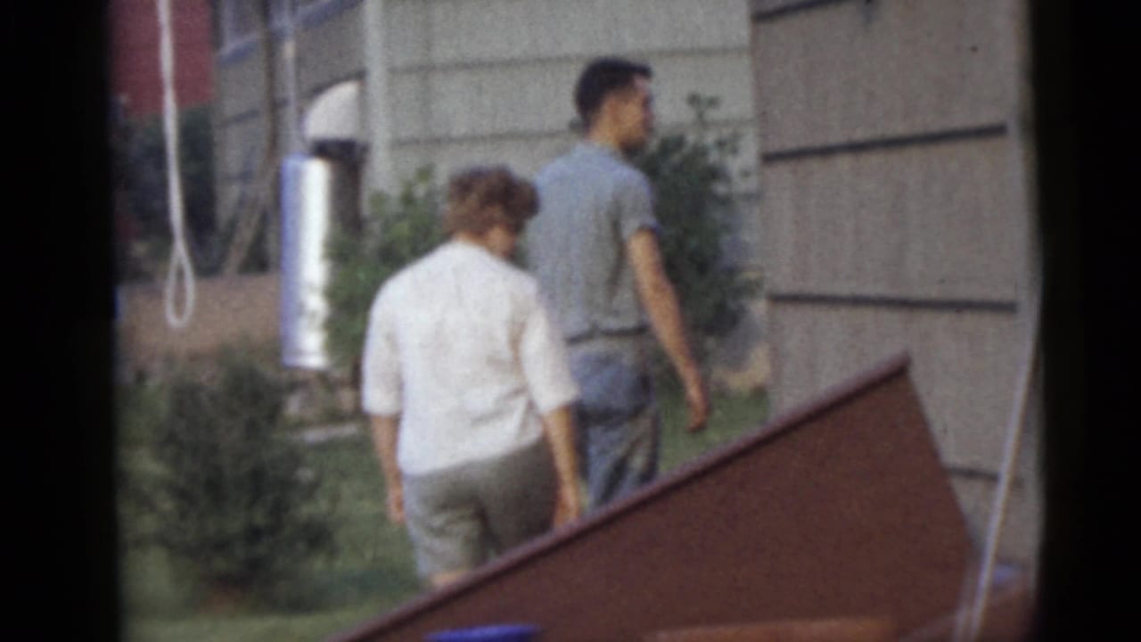 The man and woman continue walking away through a Camden backyard, passing house siding, shrubs, and a slanted wooden structure.