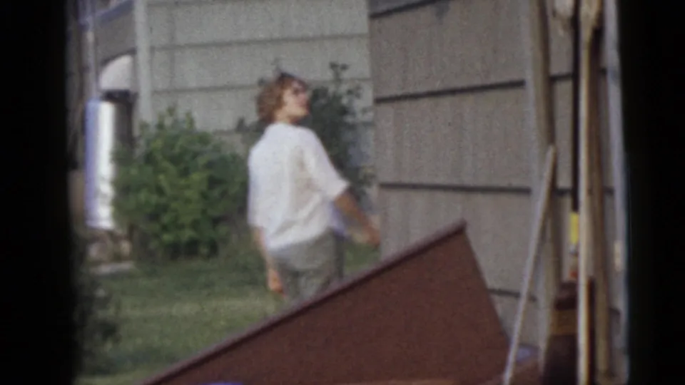 A woman in a white top walks through the 1960 Camden backyard, glancing upward near the house and greenery.