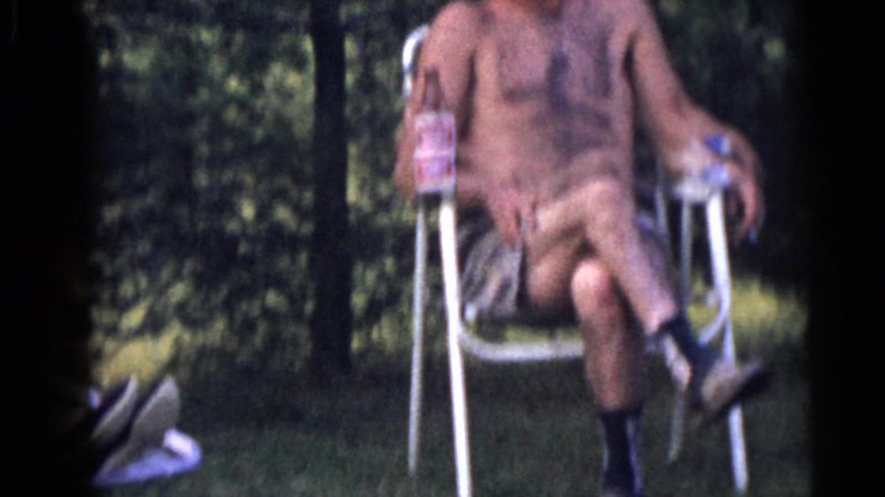 Shirtless man seated in a metal lawn chair holding a bottle outdoors.