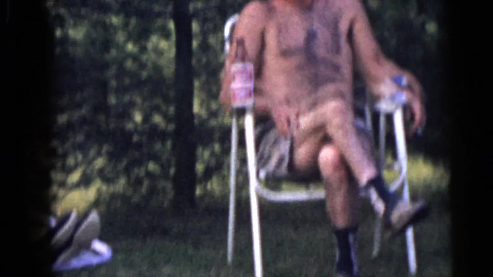 In 1960 Camden home-movie footage, a shirtless man sits in a metal lawn chair with a bottle near trees and grass.