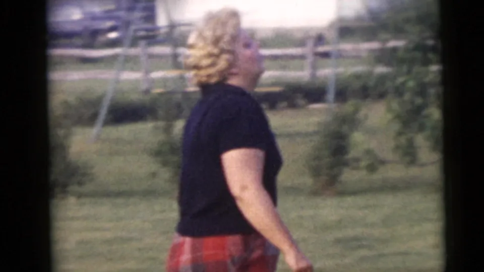 In supplied 1960 Camden home-movie footage, a woman in a black shirt and red plaid pants walks through a suburban yard.
