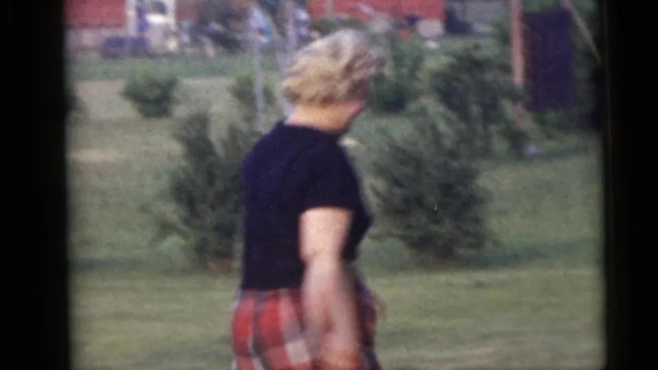 The woman continues walking past low shrubs in the residential yard, her red plaid pants and black shirt visible in soft 8mm color.